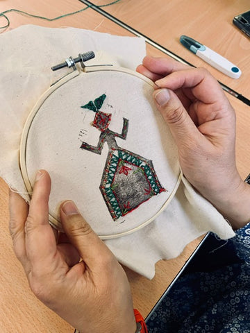 Two hands hold an embroidery hoop, the mokosh symbol is printed onto the canvas and delicate stitch work on red and green has been added.