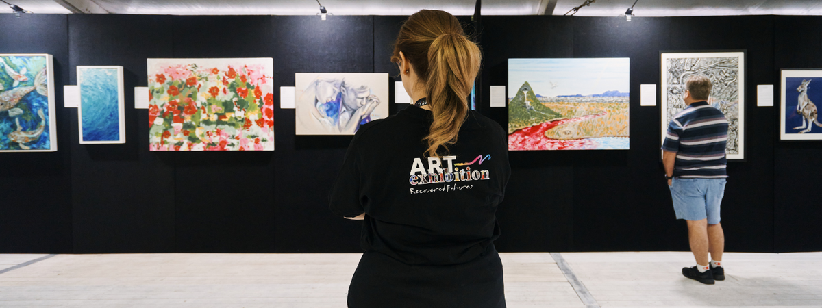 A woman with her back turned, looking at an array of artworks hung up on a black wall. The back of her shirt has a Recovered Futures Art Exhibition Logo o it.