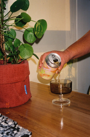 A person pouring a can of tina into a small wine glass. There is a plant next to it and the afternoon light is shining