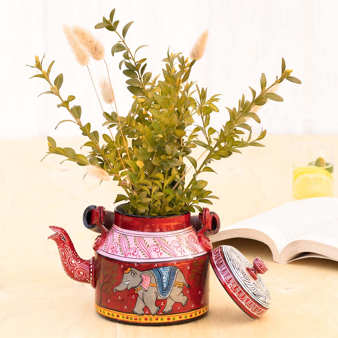 Red aluminium planter with an elephant filled with green twigs standing on a table with a open book and a glass in the background