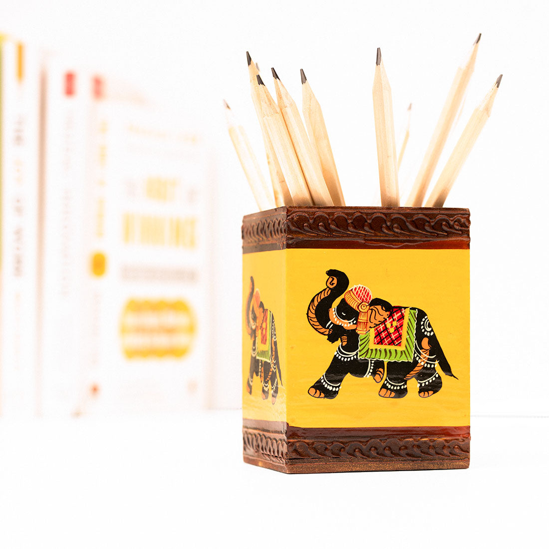 Wooden yellow pen stand with elephant motifs holding pencils placed on a white surface with a collection of books in the background.