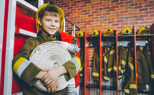 Kid dressed as a firefighter holding a hose in front of a firetruck