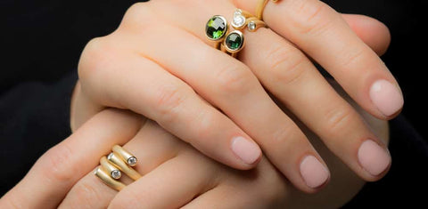 A woman's hands resting on top of each other, one wearing two gold rings with green accent gems and the other wearing a single gold ring with silver accent gems