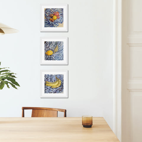 Three art prints of oil paintings by artist Harriet Lawless of bananas, tomatoes and a lemon on a blue and white stripey cloth, framed and hung on a wall. 