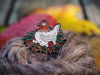An enamel pin with a cartoon chicken sitting on eggs and red balls of yarn. Her nest is made of brown and green leaves.