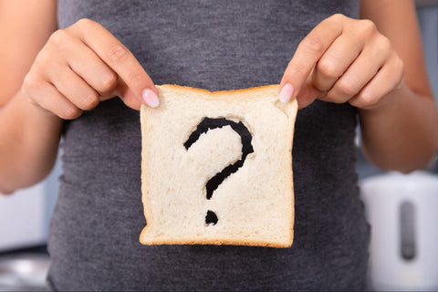 Vor ihrem Bauch hält eine Frau eine Scheibe weißes Toast, aus der etwas Teig in Form eines Fragezeichens ausgeschnitten ist