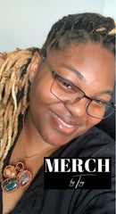 A woman with blond and black locs and black glasses smiling with a crystal necklace on. The words Merch by Toy are on top of the picture.