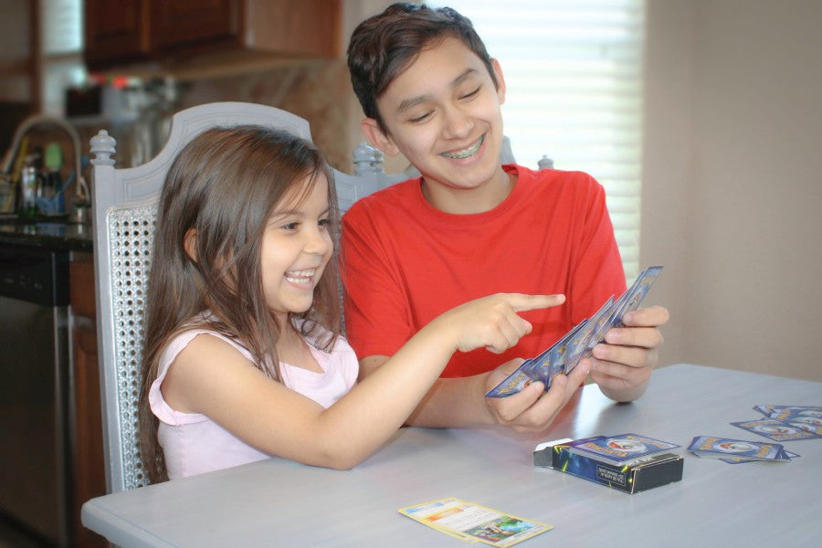 Two children indoors looking at a handful of Pokémon cards and smiling. Daytime. Dining room table.