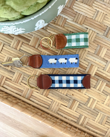 3 needlepoint key chains sit on top of a rattan table. Two are gingham in pattern and one is blue with white sheep.
