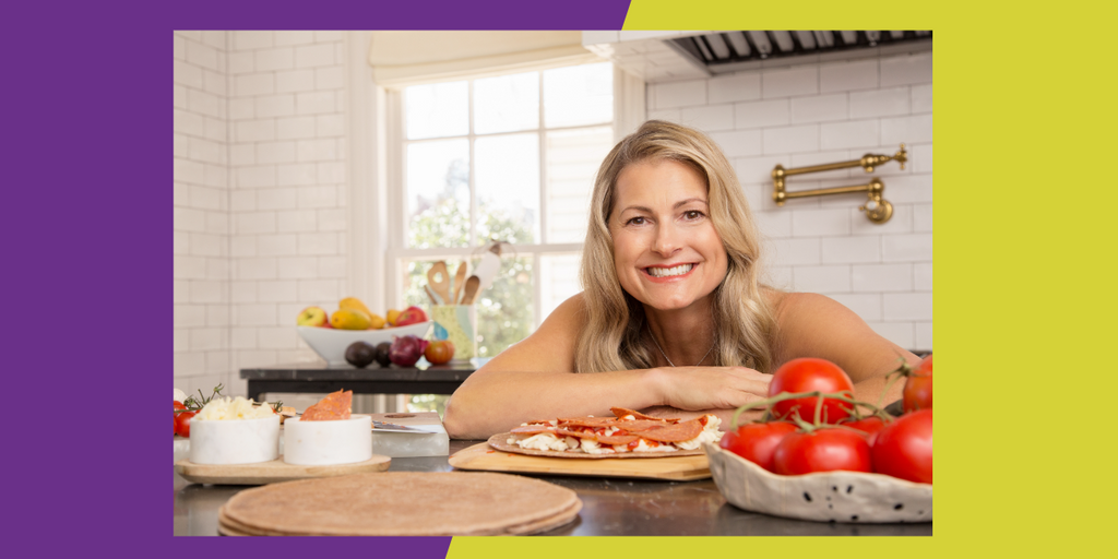 A woman smiles near a countertop with tomatoes and a pizza