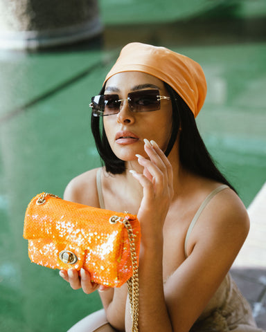 Girl holding orange sequin mini crossbody bag, with black rimless sunglasses and and orange headscarf bandana 