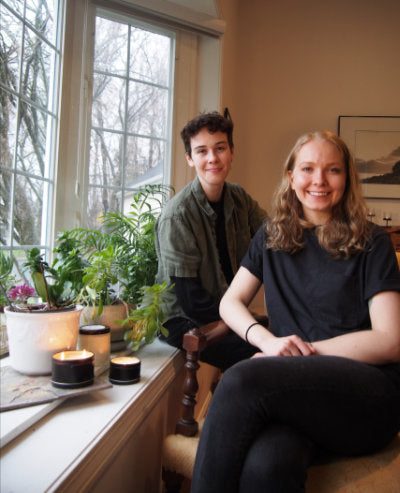 image of two people, owners of queer candle company, sitting in front of a window with samples of their candles on the surface next to them