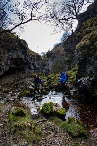 A man and woman cross a shallow river in a gorge