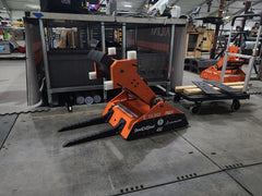 Switchback, a bright orange and black robot, sits on the ground in front of a stainless steel table. Switchback has long pointed forks attached to its front, and the arm is positioned at an angle so Switchback looks ready to fight.