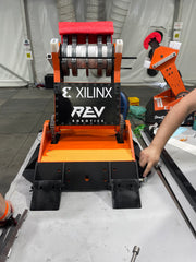 Switchback, a bright orange and black robot, sits on a stainless steel work table, facing away from the camera. The large Xilinx and REV Robotics logos are visible on the front arm plate. Switchback has steel side skirts attached to the drive train, and a set of small wedges are screwed onto the back.