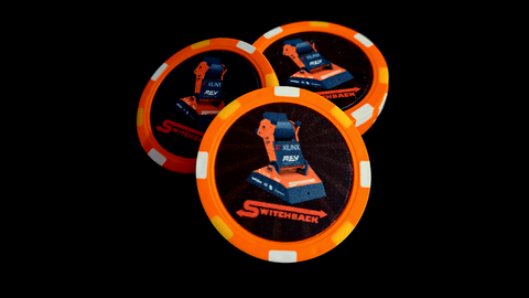 Orange poker chips on a black backgrounds. The poker chips are printed in the middle with a circular pattern and the orange and gray Switchback robot. The Switchback logo is printed right beneath the image of the robot in orange.