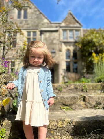 toddler girl standing infront of old cotswold stone manor house wearing cream dress and blue cardigan