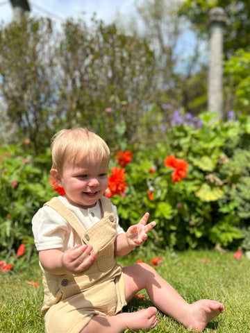 happy baby sat in sunshine infront of summer flowers wearing cream overalls