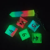 A set of five glowing six sided dice and a crystal shaped keychain in the dark. There is one die each of blue, green, yellow, and pink, and the fourth die and keychain contain layers of those colors.