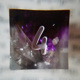A close up image of the face of a d6 die containing a dark feather and silver flakes in half clear half purple resin.