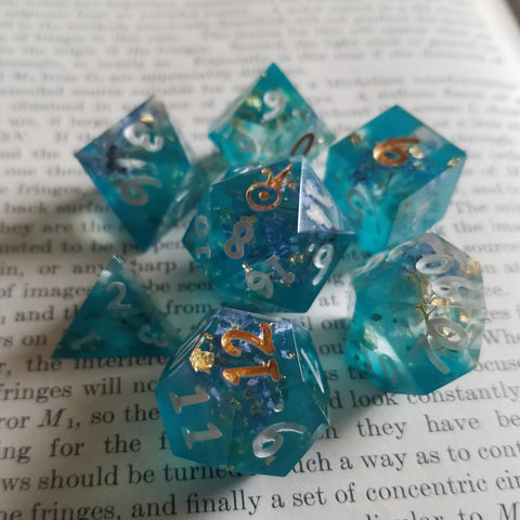 Teal dice sitting on the pages of an open book. The dice also contain forget-me-not flowers.