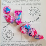 A set of pink and blue dice on top of a spiraling graph in a science book. The dice contain a lattice of white between the pink and blue clouds of resin.