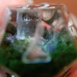 A close up image of the inside of a d12 die. It contains a headstone surrounded by dark green grass, tiny purple flowers and smoky white fog.