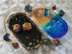 A set of jumbo dice in which every die is different, a dark colored moon-shaped necklace, and two small resin trays. One tray looks like the beach and the other is mainly dark green with a border of flowers.