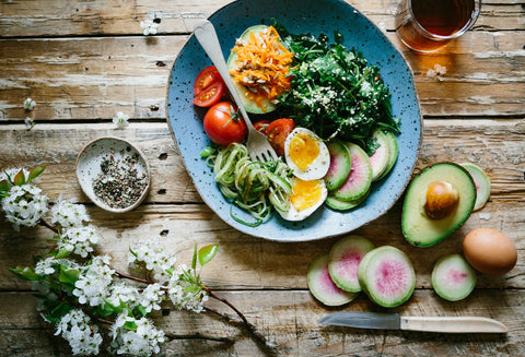 A rustic wooden table laid out with a colourful plate of mixed salad with eggs, avocado & spiralised vegetables, flanked by fresh ingredients and white blossoms.