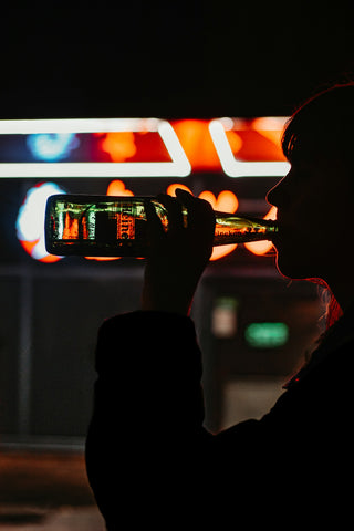 Silhouette of a person drinking from a bottle against a backdrop of neon lights at night.