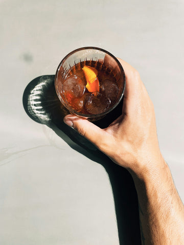 A hand holding a glass of iced drink garnished with an orange peel, casting a shadow on a sunlit surface.