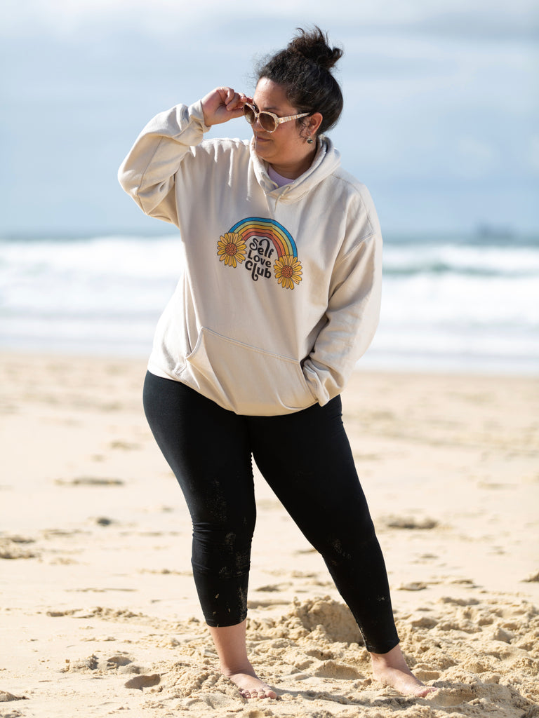 Woman at the beach wearing a sand color hoodie with Self Love Club written on it | Raiana's Vibes