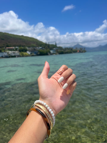 Photo of a manicure using semi cured gel nails from Iwi Nails. Hand is in front of a picturesque ocean and mountain background. Nails are orange and white with a miter shell print.