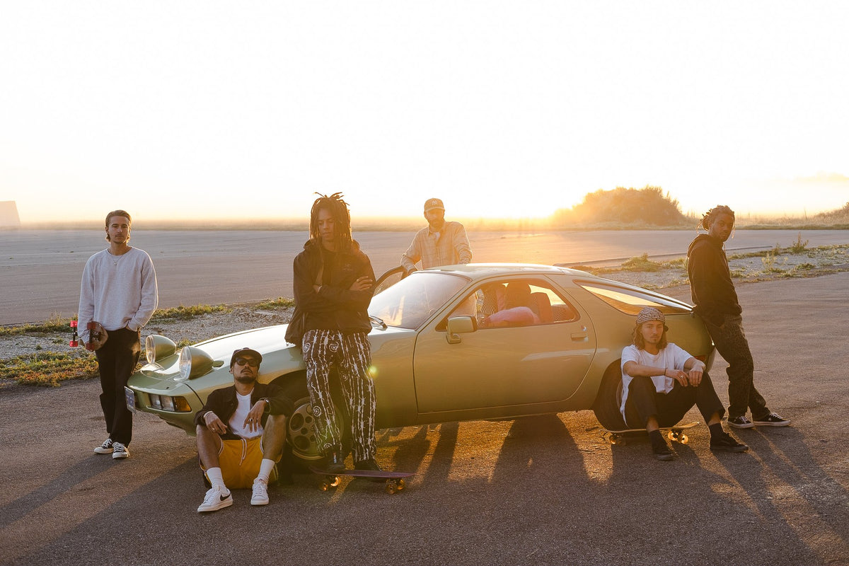 A group of skateboarders sitting and standing next to a Porsche as the sun sets