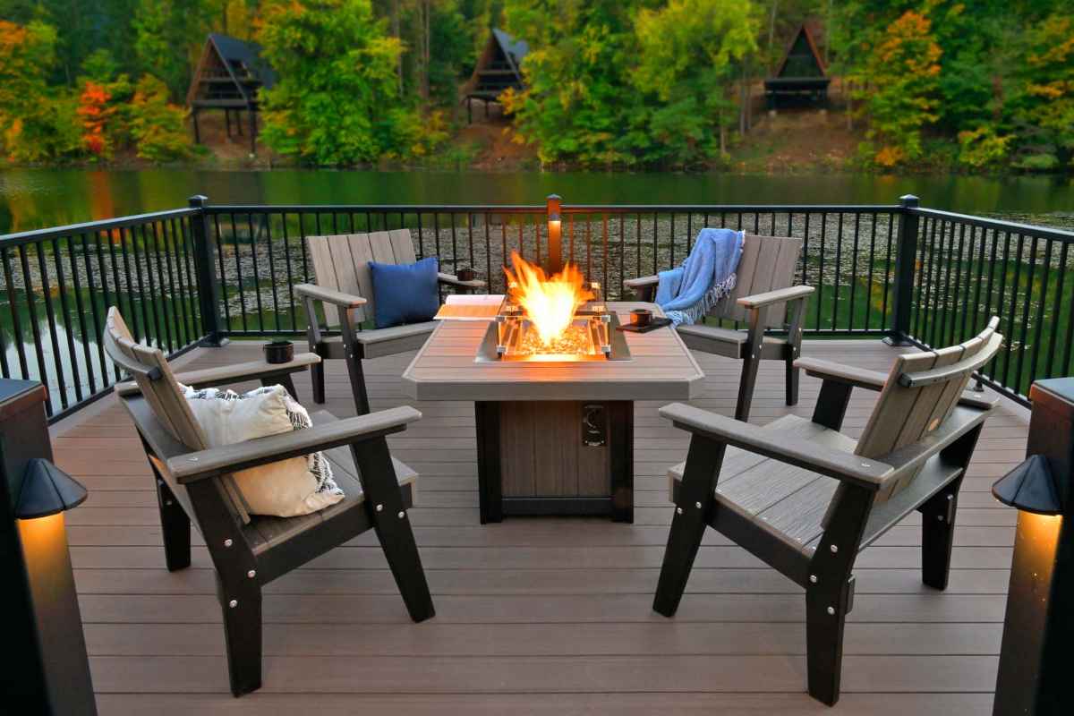 Four wooden Adirondack chairs surround a glowing firepit on a deck beside a lake, illustrating one of the 8 Trends in Outdoor Living Spaces that emphasize warmth and gathering spots