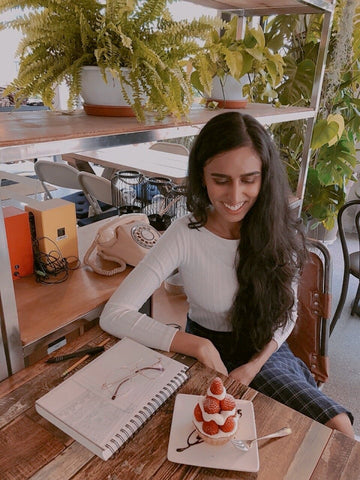Portrait of Sabbie Narwal, the artist behind The Paper Narwhal brand. Image features the artist sitting at a table and looking down at a pastry and sketchbook.