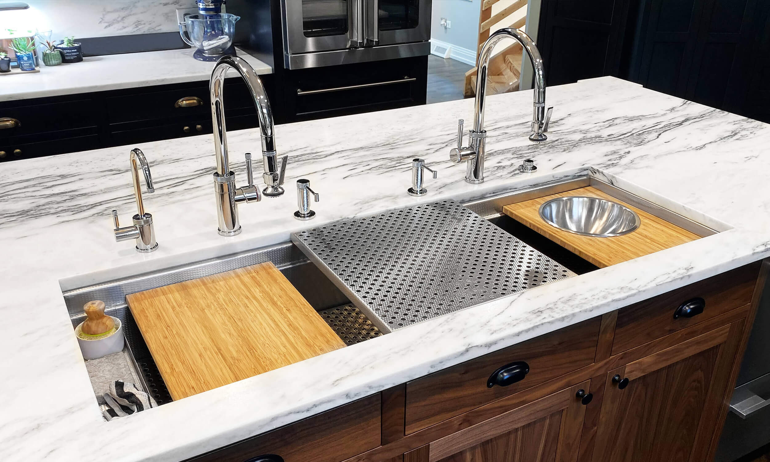 Dual Tier Workstation Sink in Prestige Stainless Steel. USA Made by Havens Luxury Metals. Kitchen has white marble countertops and dark wood cabinetry.