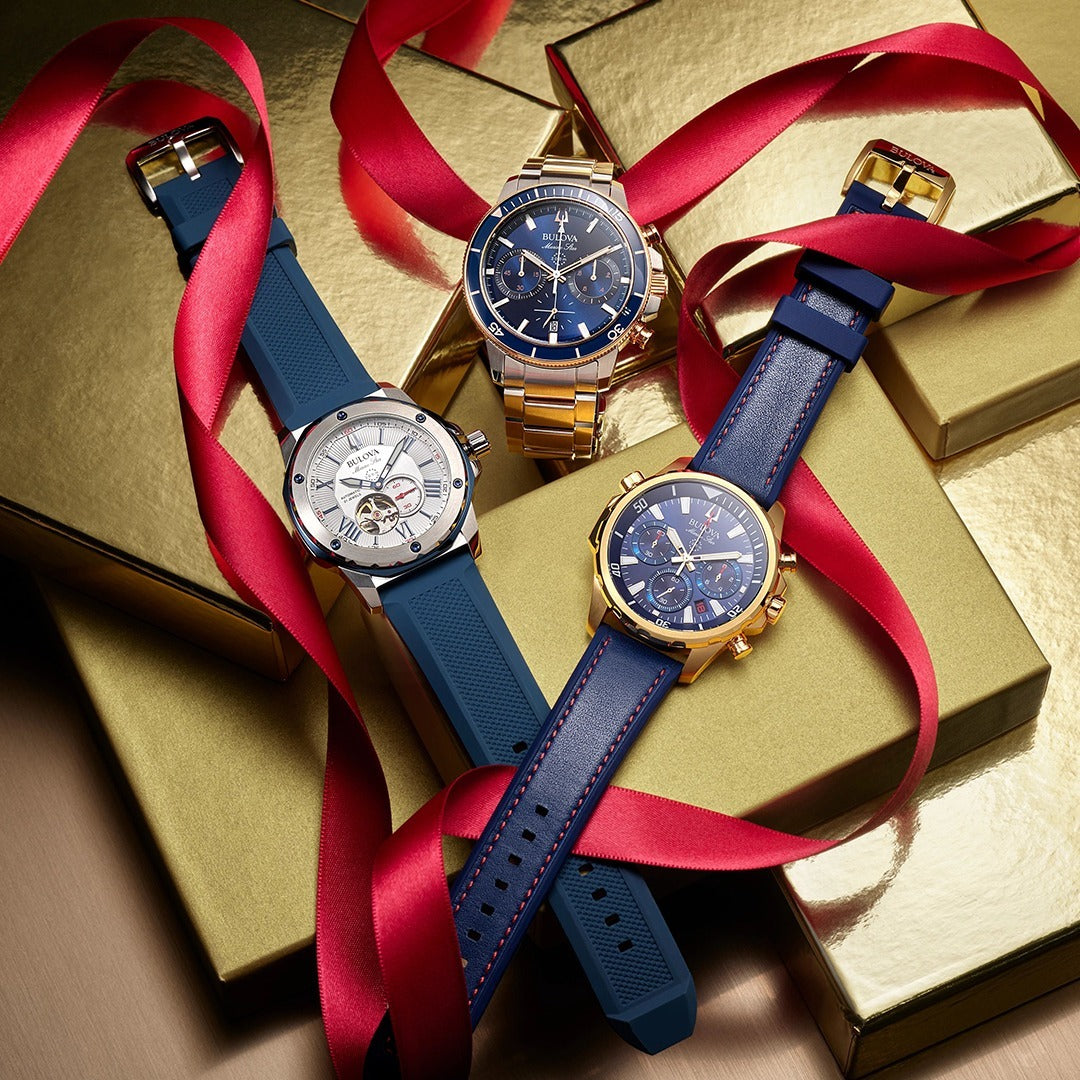 Three watches are stacked on a pile of gold colored boxes with red lace wrapped around the watches