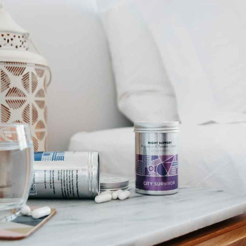 City Survivor magnesium supplements in metal containers with a purple label stand on a marble bedside table next to a fresh white bed. A glass of water and some loose capsules stand beside the container