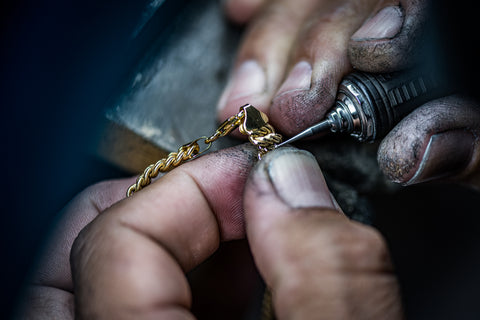 Up close photograph of a mans weathered hands working on a gold bracelet with tools.