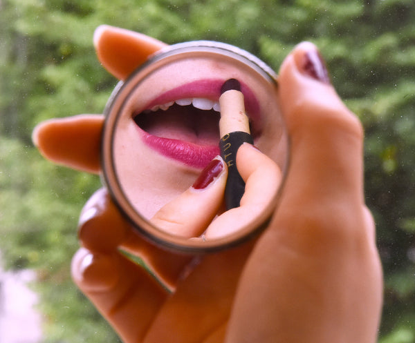 Reflection of woman applying Vermouth Lip crayon with trees in background