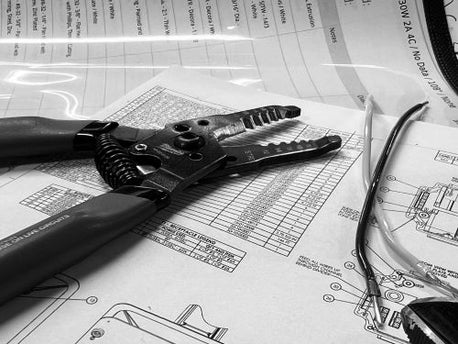 Black and White image of wire strippers and cut wires laying on top of a engineering drawing and a bill of material
