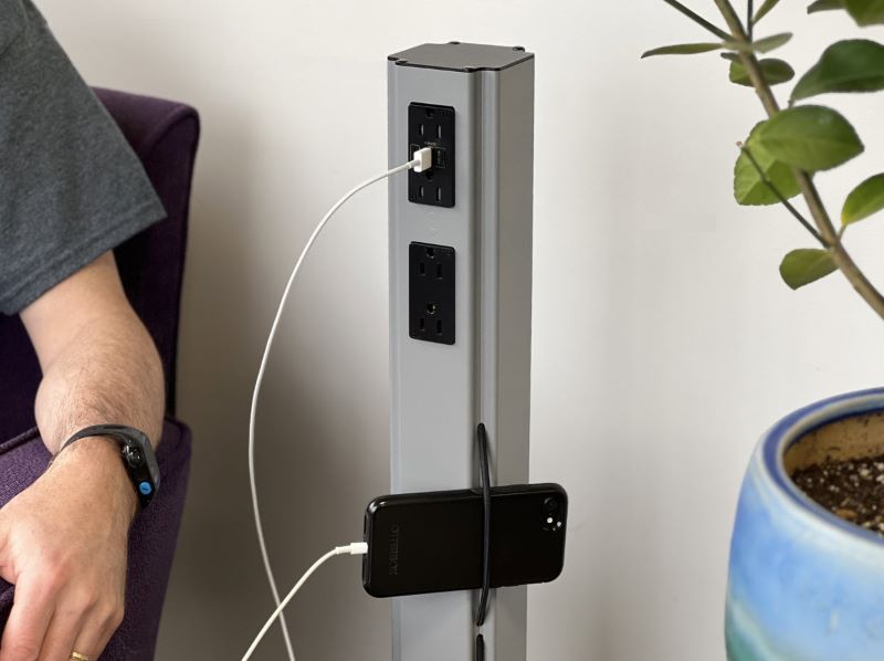 Gray MG Power Plus shown charging an iPhone. A man's arm is visible as it rests on the arm of a chair. There is also a partial view of a plant on the right of the MG Power Plus