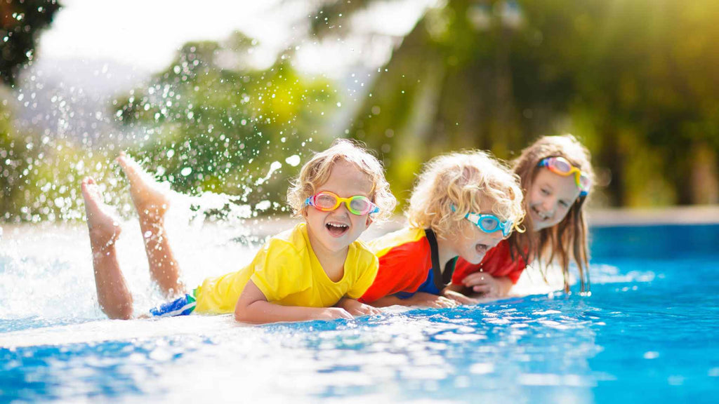 Three children with goggles playfully splashing in a sunlit swimming pool, enjoying an afternoon.