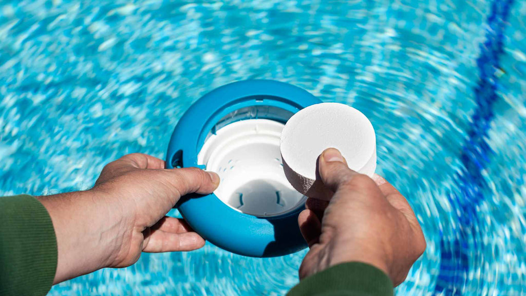 Hands placing a chlorine tablet into a floating dispenser in a swimming pool with rippling blue water.