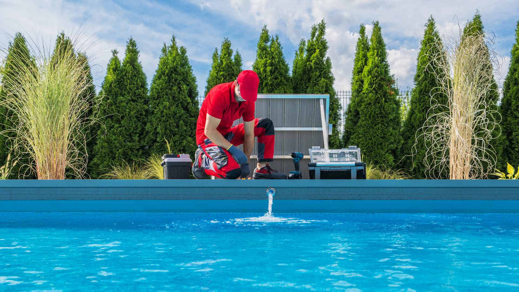 A person in a red outfit kneeling by a swimming pool, examining equipment on a sunny day with greenery in the background.