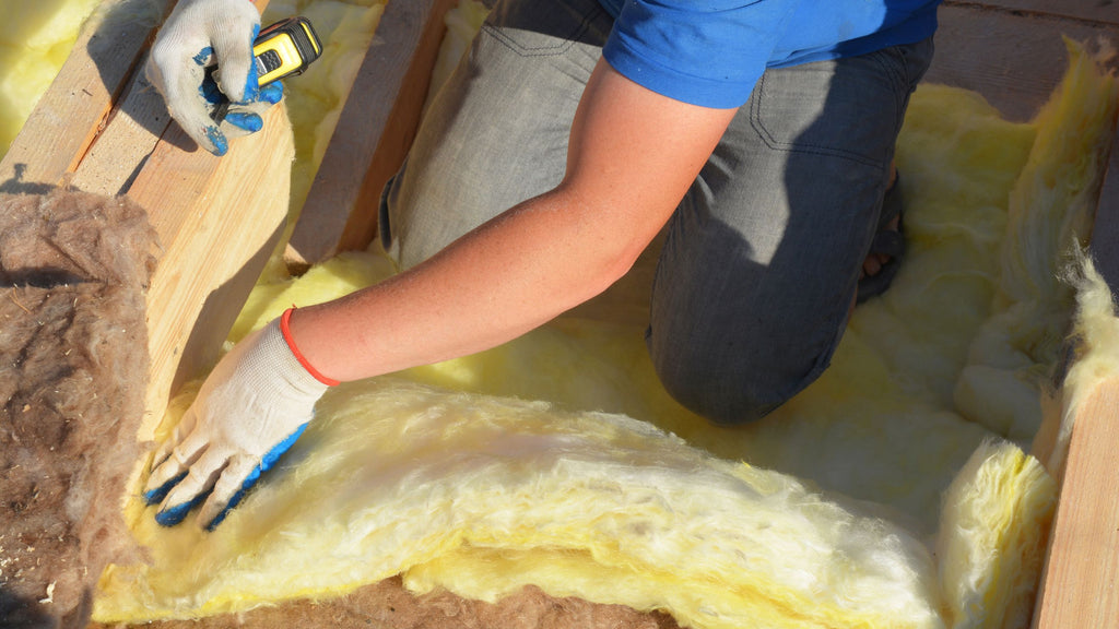 A man is laying insulation on a wooden floor, unaware of the potential fiberglass irritation to his skin.