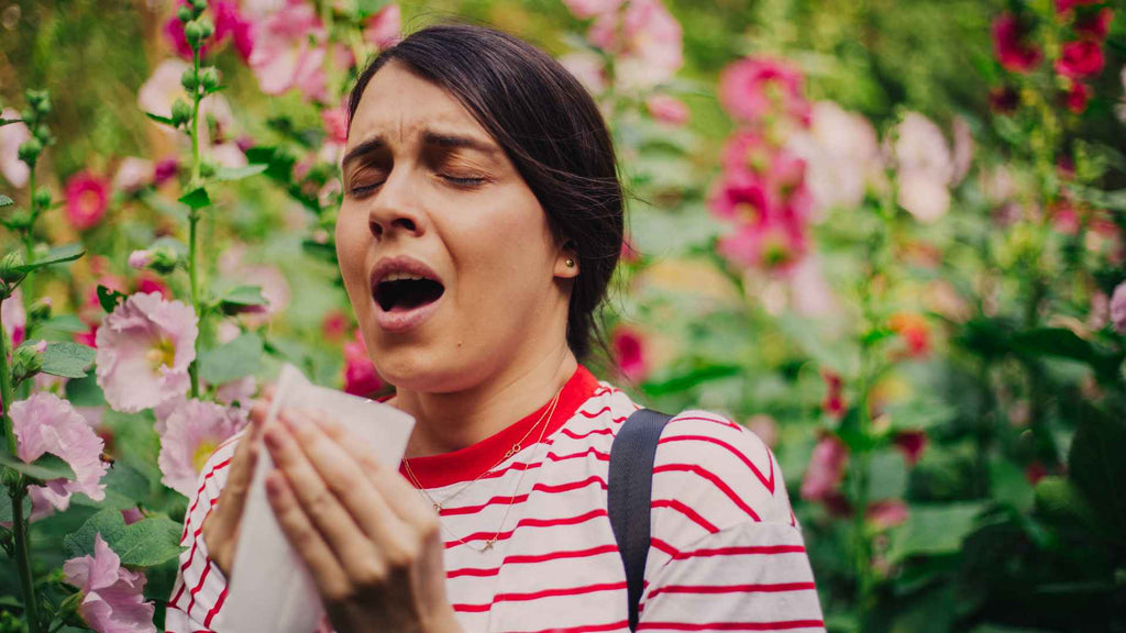 A woman with hay fever blowing her nose in a garden full of flowers.