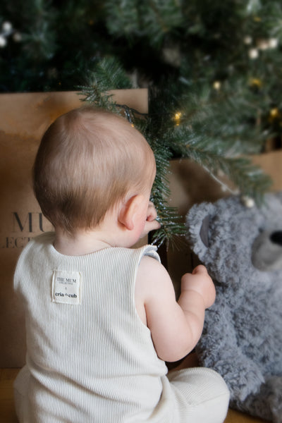 A baby sat by a christmas tree wearing a The Mum Collective romper