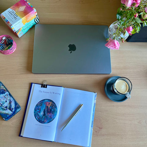 journal opened on desk, with pen lying on top. Other items on desk: phone cover, deck of tarot cards, Apple MacBook, vase of roses and a freshly brewed espresso